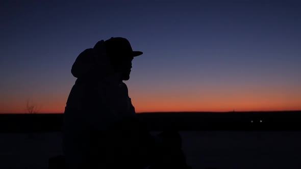 A young man in a cap sits thoughtfully at sunset and dreams