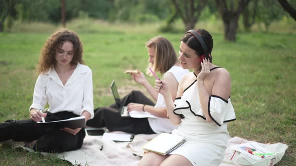 Young Woman Enjoying Music in Headphones Sitting on Summer Spring Lawn in Park with Colleagues