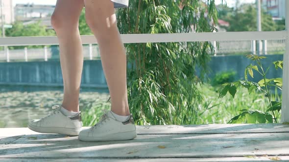 the girl's feet in white sneakers are on white wooden boards against the background of a green willo