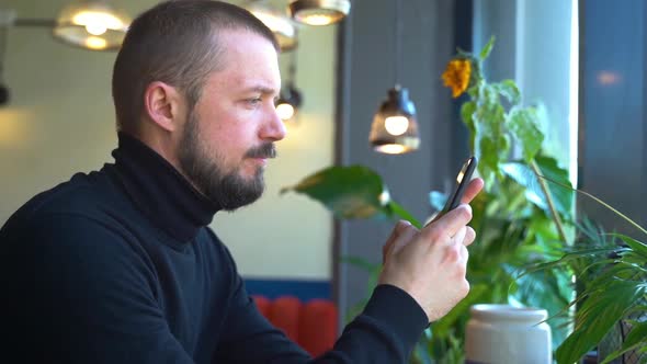 American Bearded Man Is Using Phone and Typing Message at Table in Cafe Spbd.