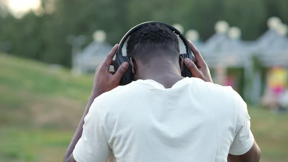 Black Man Putting on Headphones Walks Against Blurry Park