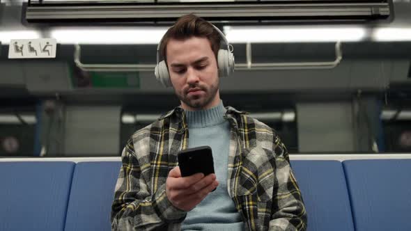 Focused Young Man Rides the Subway Listens to Music with Wireless Headphones and Uses a Smartphone