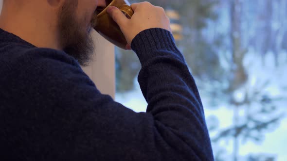 Closeup of a Brutal Man with a Beard Stands at the Window and Looks Into the Distance Drinking Hot