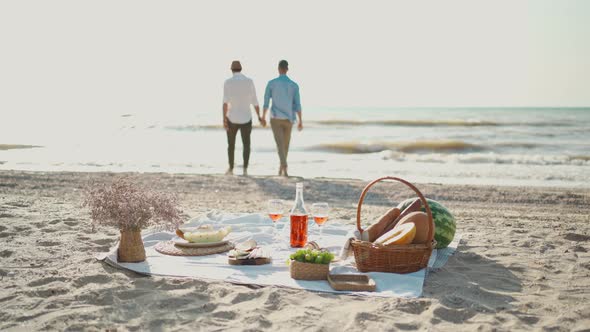 Silhouette Gay Couple Walking By Sea Beach Focus on Picnic Blanket with Wine Glasses and Food