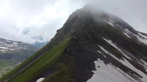 Drone shot (rotating) of a Dark Mountain, partially covered in snow and in the Clouds