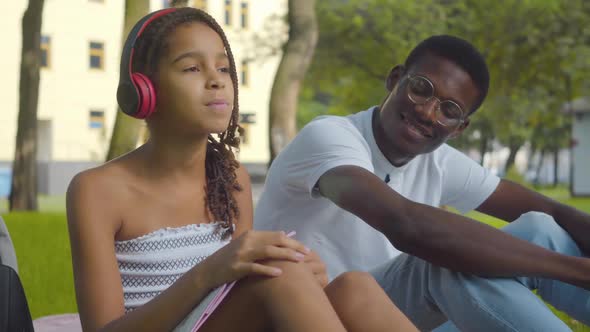 Portrait of Cheerful Cute Girl Listening To Music in Headphones and Giving Earphones To Young