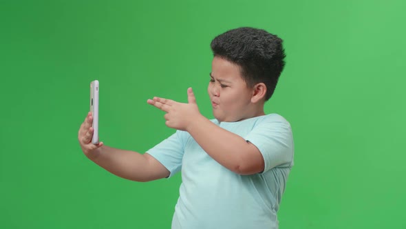 The Smiling Asian Little Boy Making Selfie While Standing On Green Screen In The Studio