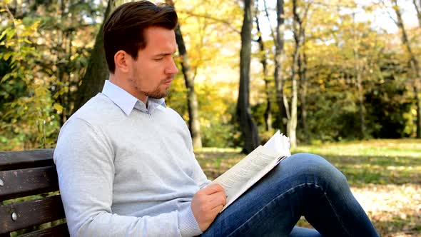 Young Man Sits on Bench in a Park and with Interest Reads a Book