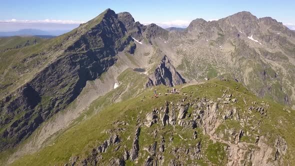 Wide aerial view of climbers on mountain summit with green grass and bright blue sky. Jagged mountai