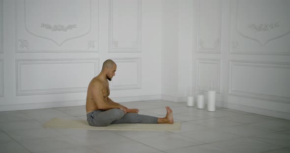 Male Yogi Is Sitting on Sportive Mat on Tiled Floor in Hall with White Walls, Stretching His Legs