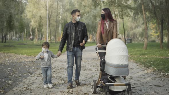 Wide Shot of Friendly Young Family in Covid Face Masks Walking in Spring or Summer Park and Talking