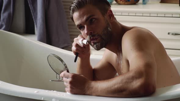 Handsome Young Bearded Man Shaving Beard in Bathrobe in Bathroom.