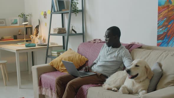 Black Man Using Laptop and Petting Dog on Sofa