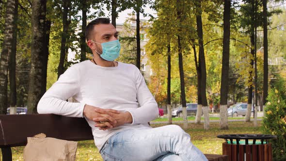 A Handsome Businesslike Young Man Sits on a Bench Wearing a Medical Mask