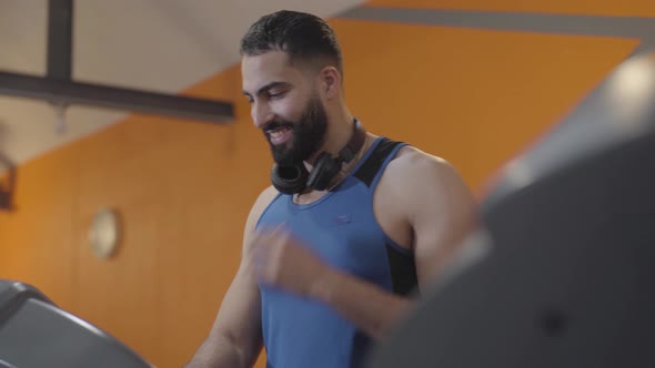 Happy Middle Eastern Man Adjusting Treadmill in Gym, Putting on Headphones and Training