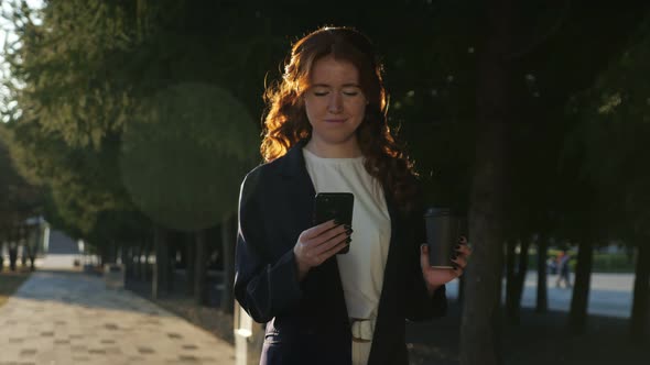 Positive Young Woman Holds Cup and Mobile Phone in City Park