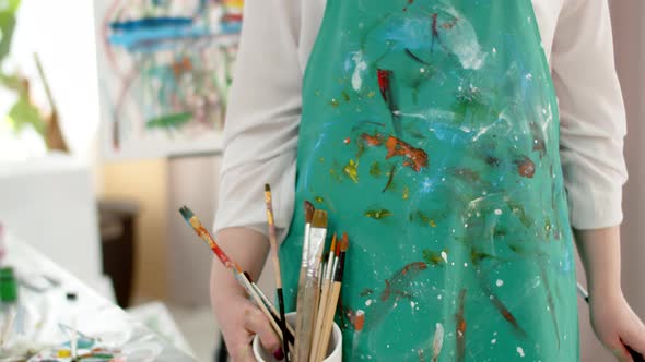 Woman artist in paint stained apron stands in workshop and holds brushes