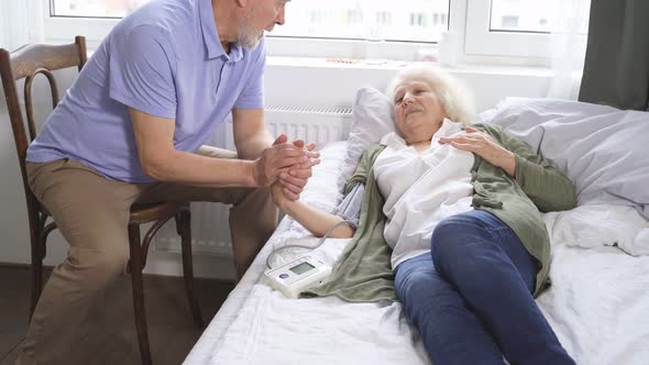 Caring Elderly Man Checks His Wife's Blood Pressure with Hypertension While Lying at Home in Bed