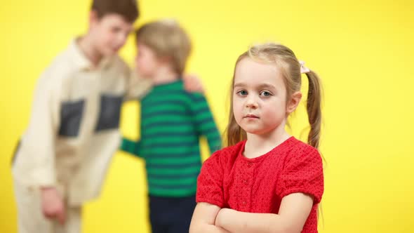Sad Charming Little Girl with Crossed Hands Looking at Camera As Blurred Boys Mocking at Yellow