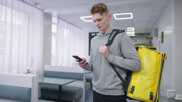 Concentrated Young Man Standing in Cafe with Yellow Food Delivery Bag Checking Online Orders in