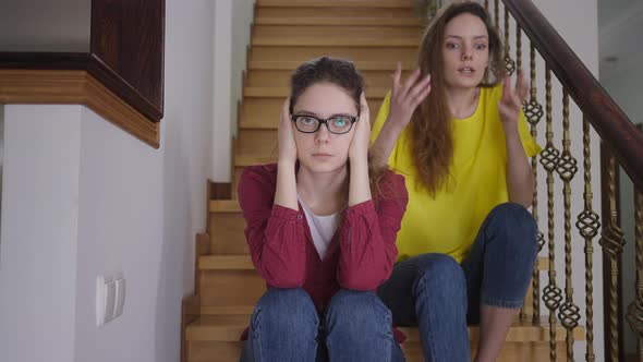 Tired Woman in Eyeglasses Covering Ears with Hands Sighing As Excited Twin Sister Talking Sitting on