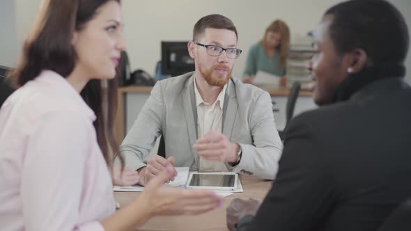 Portrait of Redhead Caucasian Salesman in Eyeglasses Talking To Excited Multiracial Couple in