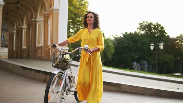 Smiling Young Girl in a Long Yellow Dress Walking Holding Her City Bicycle's Handlebar with Flowers