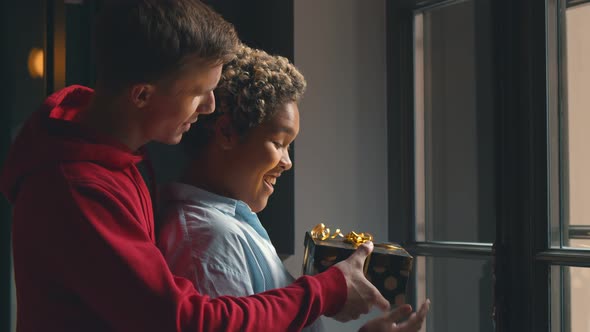 Boyfriend Giving Wrapped Box To Girlfriend Celebrating Holiday Together Standing Near Window