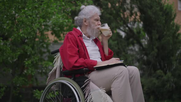 Handicapped Old Caucasian Bearded Man Drinking Cappuccino Writing in Diary with Pen Outdoors