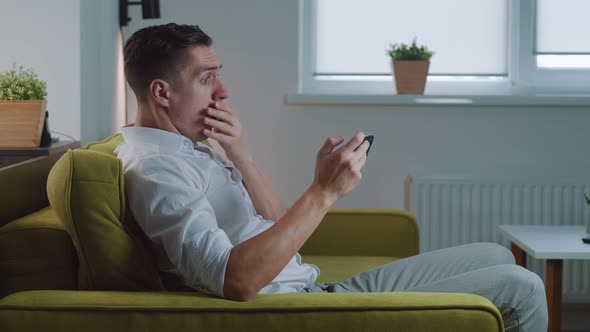 Shocked Man Reacting Message on Smartphone Covers His Mouth in Fright Sitting on Sofa at Home Living