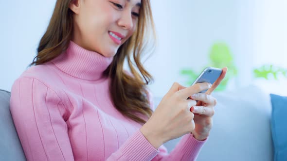 Asian young beautiful woman sitting on sofa and chatting on mobile phone in living room in house.