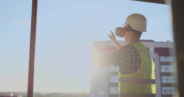 Engineer Builder on the Roof of the Building at Sunset Stands in VR Glasses and Moves His Hands