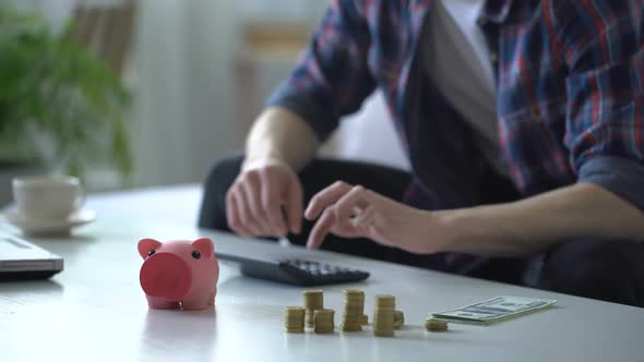 Man Putting Money Into Piggy Bank for Vacation, Counts Expenses on Calculator
