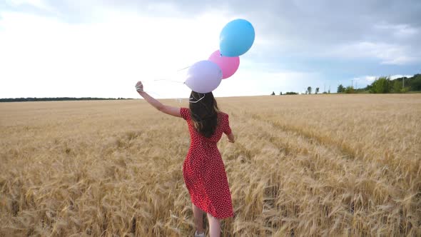 Happy Girl in Red Dress Running Through Golden Wheat Field with Balloons in Hand at Overcast Day