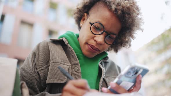 Serious African woman wearing green hoodie writing notes in notepad from smartphone
