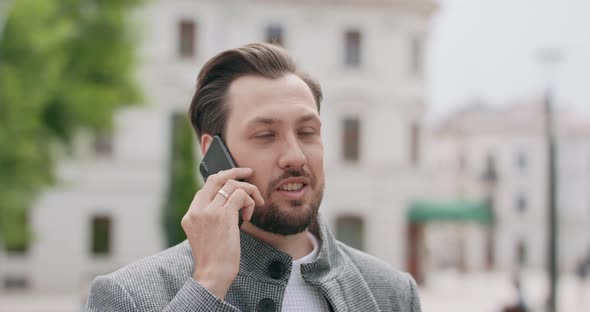 Young Man with Mustaches and a Beard is Talking on the Smartphone on the Square Buildings on