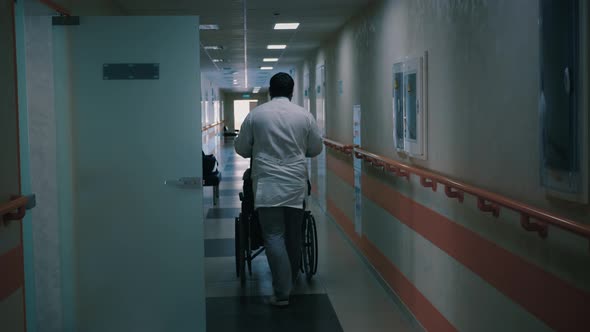 Nurse Pushing Wheelchair with Disabled Patient Through Dark Hospital Corridor