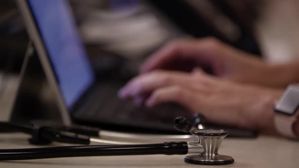 Rack focus of stethoscope on desktop to hands are typing on laptop