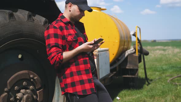 Young Attractive Farmer with Phone Standing in Field, Tractor Working in Green Field in Background
