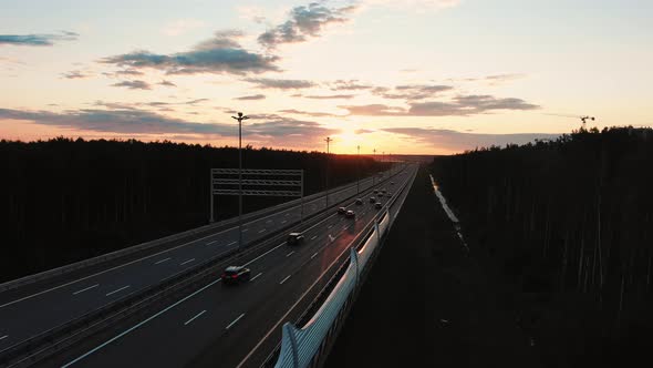 Wide Road Among Forests and Driving Cars at Orange Sunset