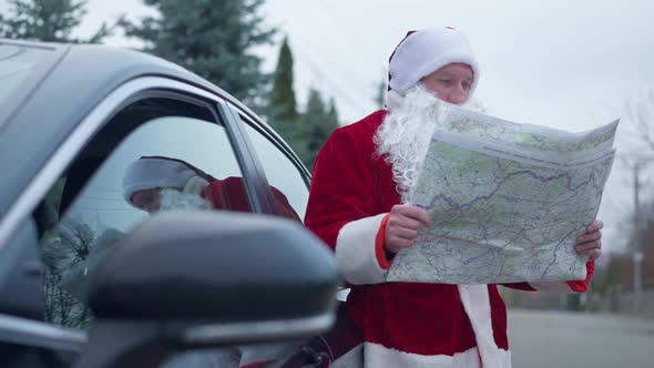 Thoughtful Man in Red Santa Costume Standing at Car with Map Looking for Direction