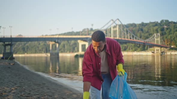 African American Man Walks Along River Beach Gathering Trash