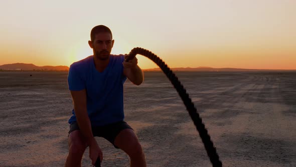 Athletic man working out with battle ropes on a dry lake at sunset