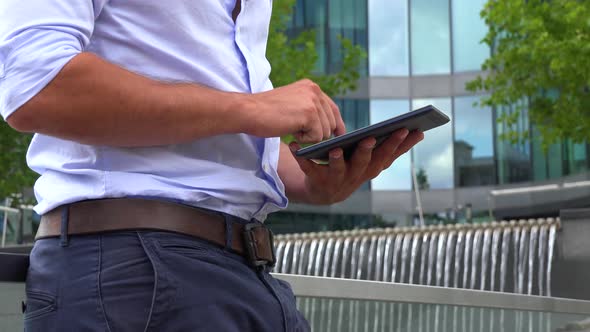 A Businessman Works on a Tablet - Closeup - an Office Building in the Blurry Background