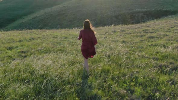 Aerial and Rear View of a Young Woman in a Red Dress Walking Barefoot Along the Hillside in High