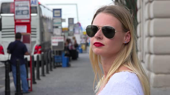 A Young Beautiful Woman in Sunglasses Looks Around a Bus Station Platform - Closeup