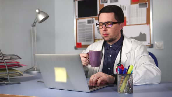 Doctor in White Lab Coat Is Drinking Tea in the Workplace and Looking at Laptop