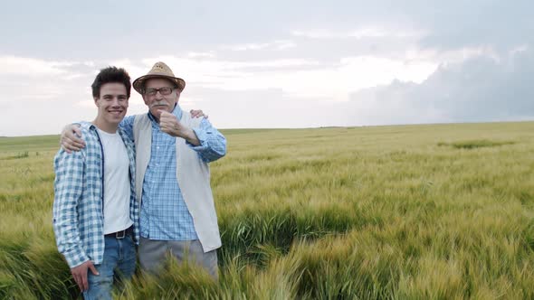 Happy Senior Farmer Embraces Young Son and They Smile at Camera in Barley Field