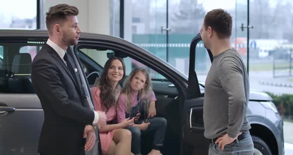 Successful Caucasian Man Talking with Car Dealer As His Wife and Daughter Sitting in Salon at the