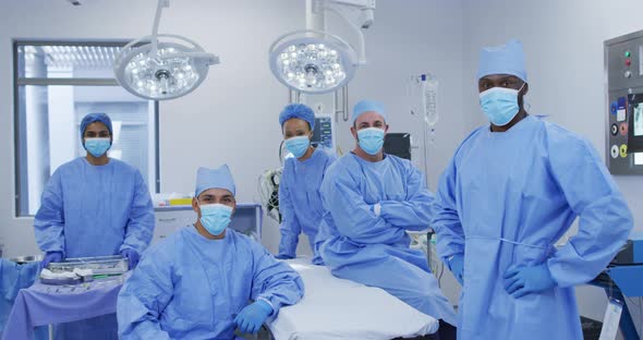 Diverse male and female doctors wearing face masks standing in operating theatre smiling to camera